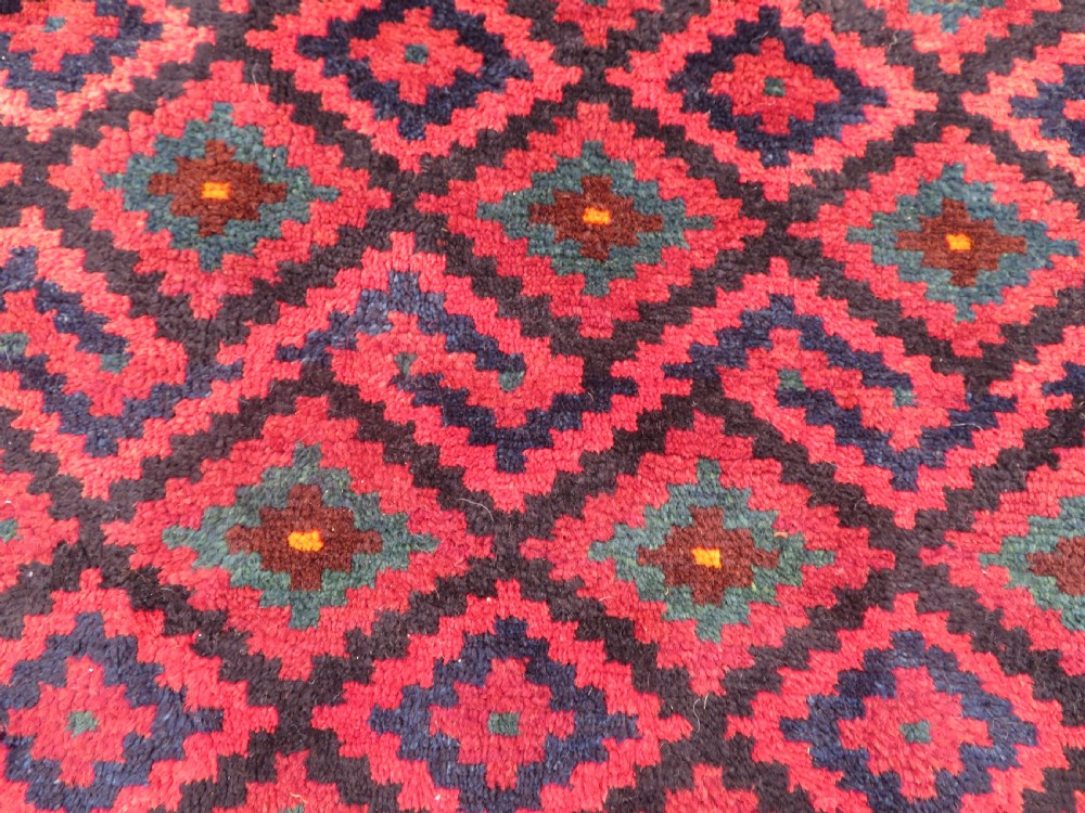 A square antique hand-knotted wool saddlebag face featuring a repeating geometric diamond pattern in red, navy blue, teal, and orange tones with light-coloured fringes. View II
