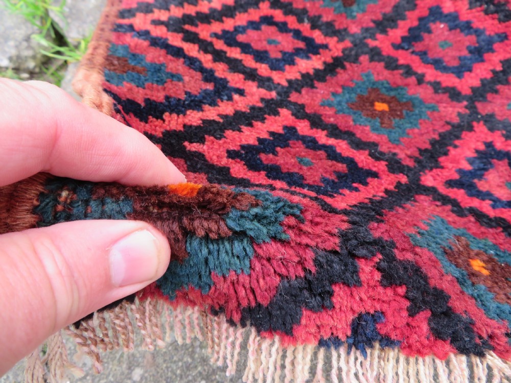 A square antique hand-knotted wool saddlebag face featuring a repeating geometric diamond pattern in red, navy blue, teal, and orange tones with light-coloured fringes. View IV