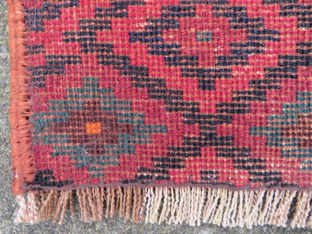 A square antique hand-knotted wool saddlebag face featuring a repeating geometric diamond pattern in red, navy blue, teal, and orange tones with light-coloured fringes. View V
