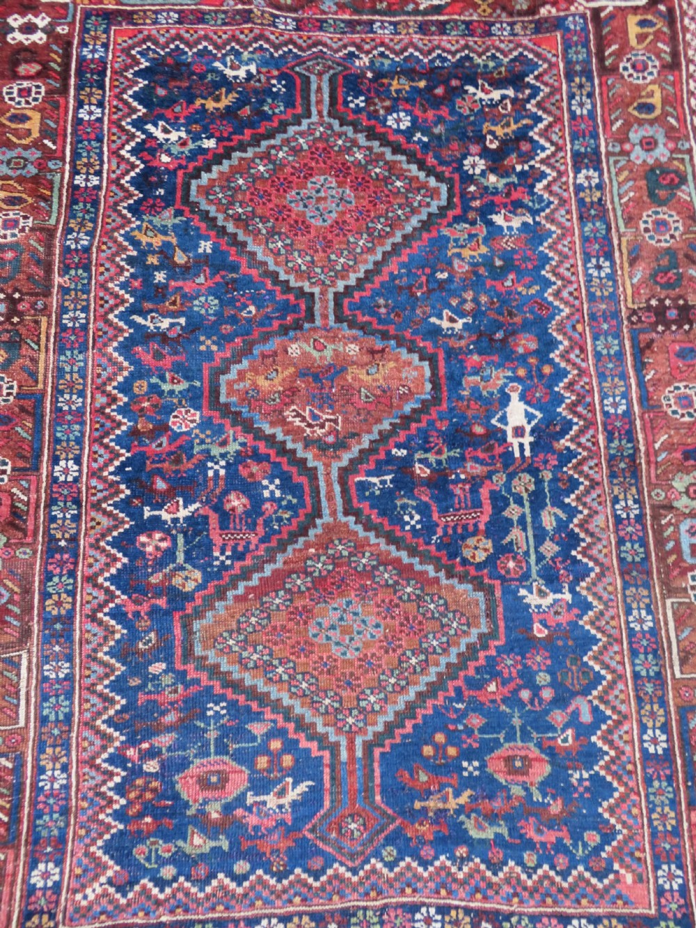 An antique Persian Khamseh rug featuring three central diamond medallions surrounded by small animal motifs and a geometric border in indigo blue and terracotta red tones. View III