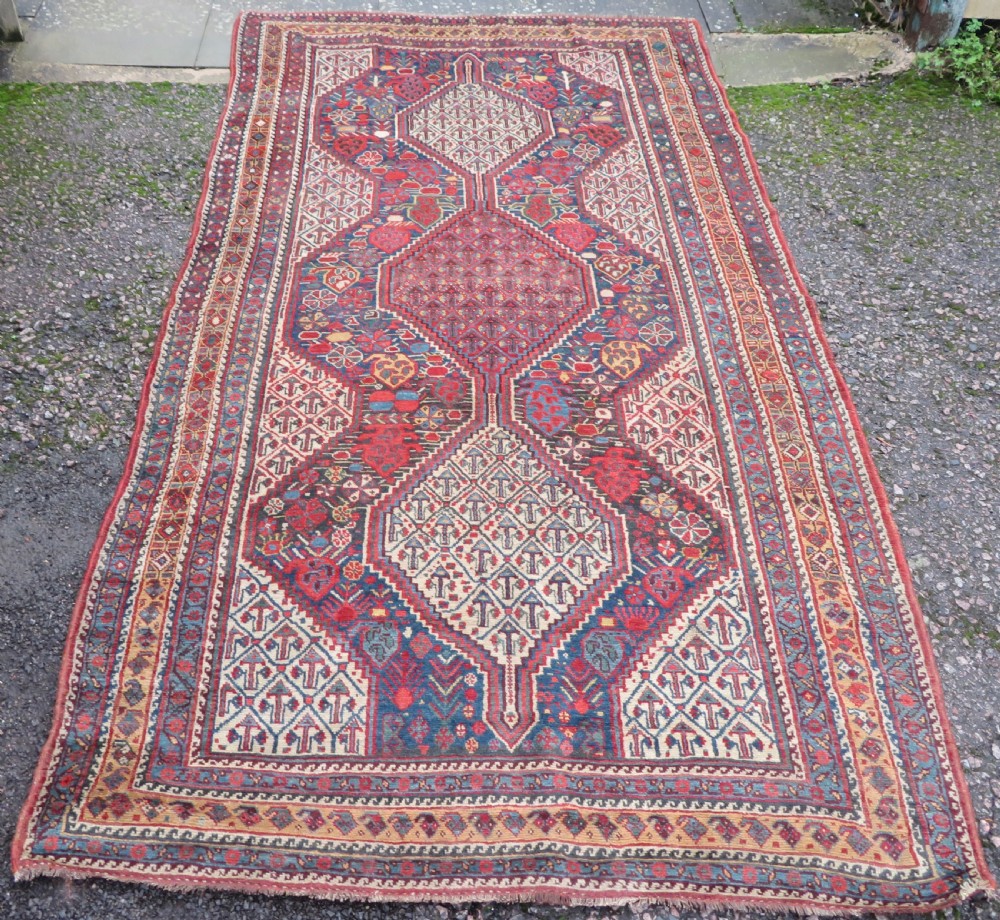 An antique Khamseh Basseri hand-knotted wool rug featuring geometric diamond medallions in red, blue, and ivory tones.