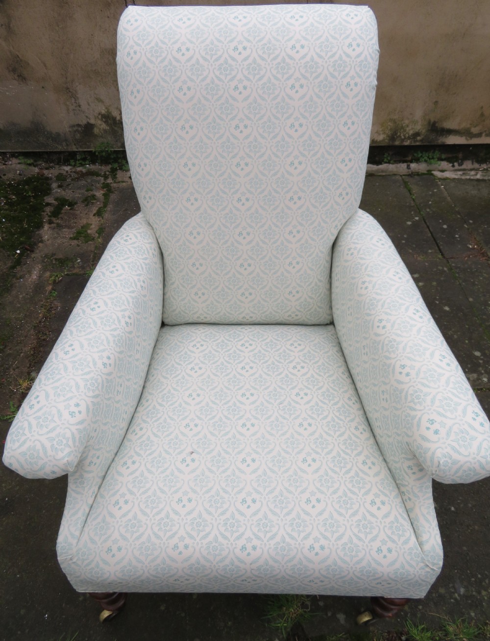 A low-seated Victorian easy chair upholstered in pale blue and cream patterned fabric with turned wooden front legs and brass castors. View VI