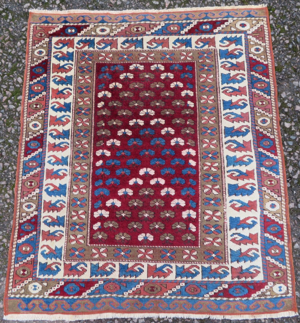 A small antique Turkish Bergama wool rug featuring a deep red central field with repeating floral-like motifs surrounded by geometric cream and blue borders.