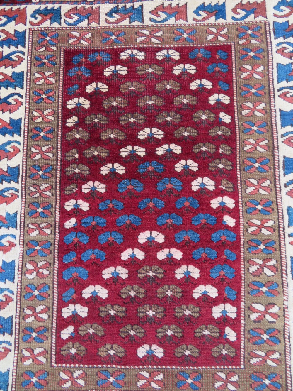 A small antique Turkish Bergama wool rug featuring a deep red central field with repeating floral-like motifs surrounded by geometric cream and blue borders. View III