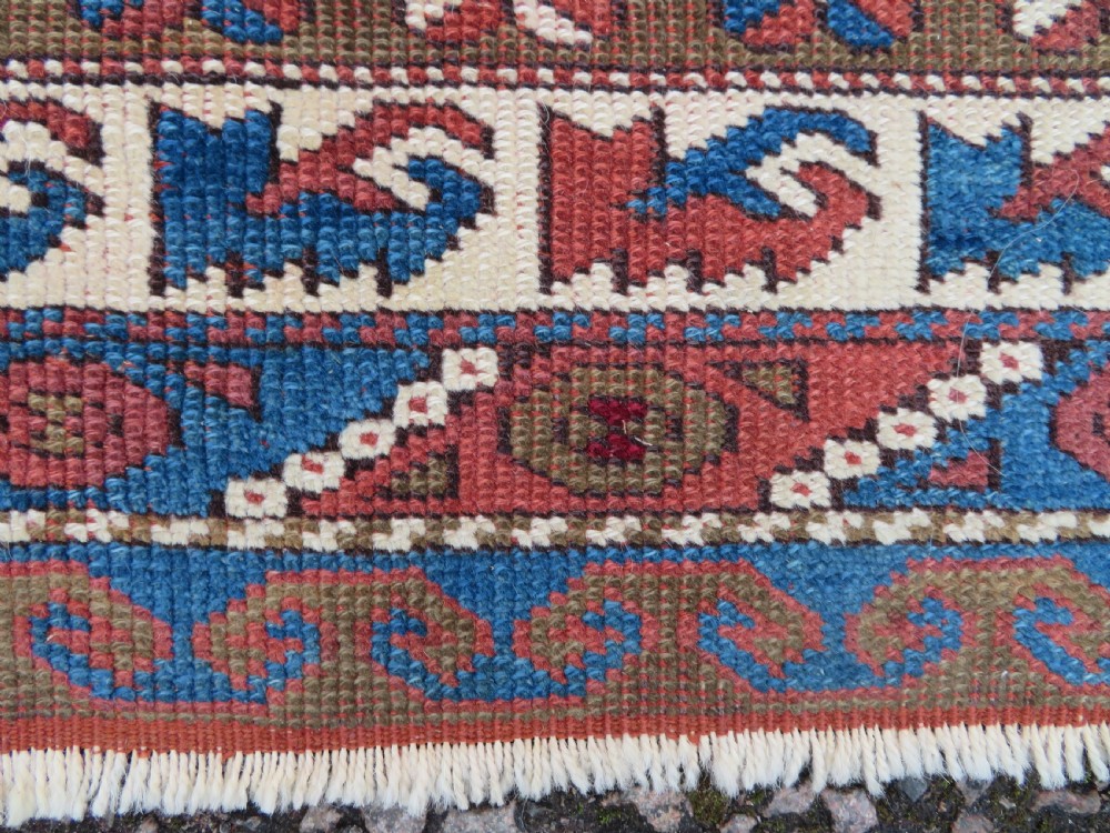 A small antique Turkish Bergama wool rug featuring a deep red central field with repeating floral-like motifs surrounded by geometric cream and blue borders. View V