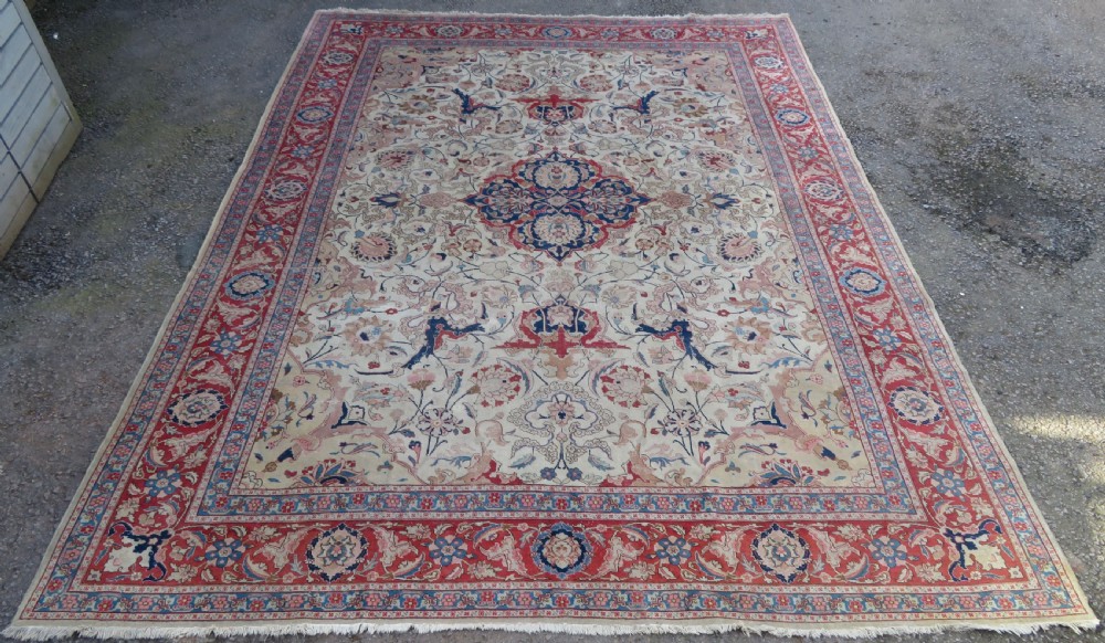 A large hand-knotted Persian Tabriz carpet featuring a central navy blue and red medallion on a cream field with floral vines and a prominent red border.