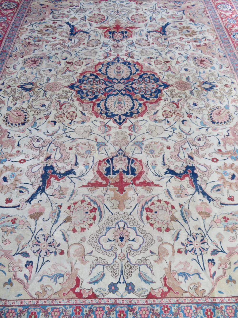 A large hand-knotted Persian Tabriz carpet featuring a central navy blue and red medallion on a cream field with floral vines and a prominent red border. View IV