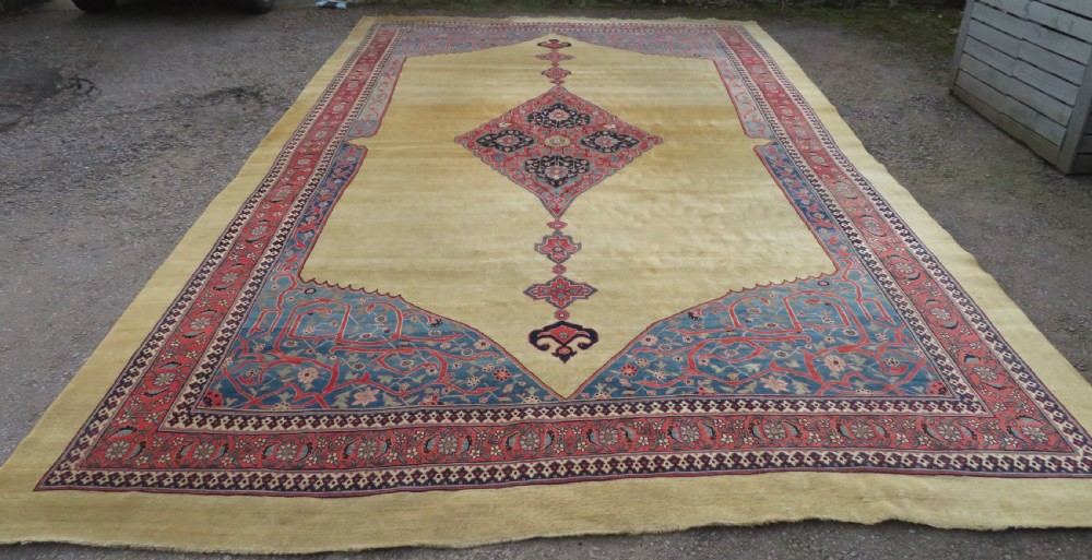 A large hand-knotted antique Persian Bidjar carpet featuring a gold yellow field, central diamond medallion, blue corner spandrels, and a patterned red border.