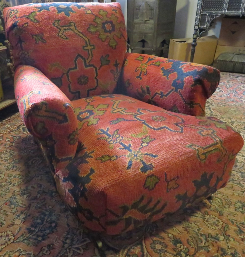 A low-slung, deep-seated Howard and Sons club armchair upholstered in a terracotta red and multi-coloured Ushak carpet pattern with turned legs on brass castors. View II
