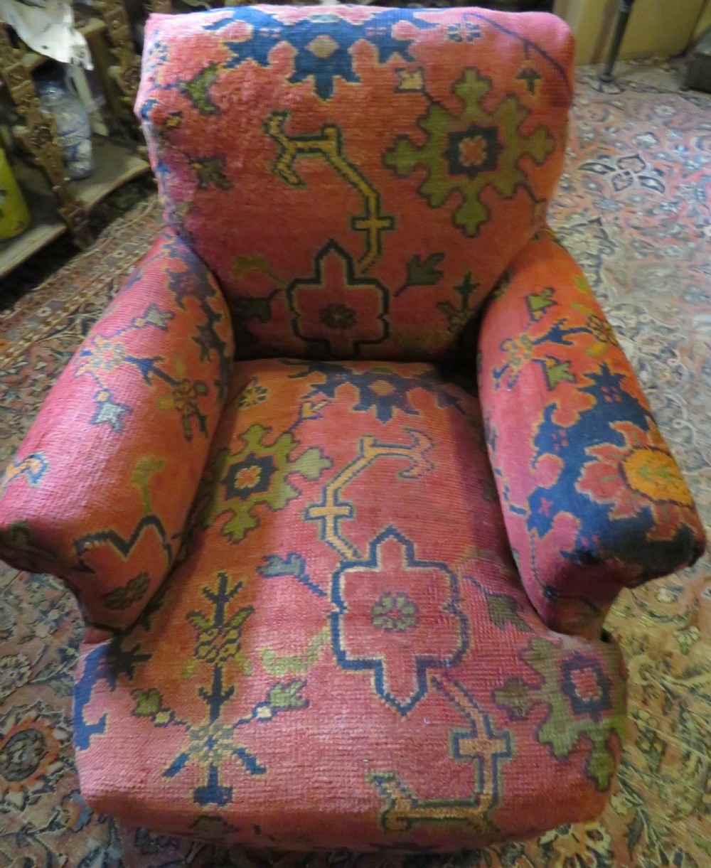 A low-slung, deep-seated Howard and Sons club armchair upholstered in a terracotta red and multi-coloured Ushak carpet pattern with turned legs on brass castors. View III