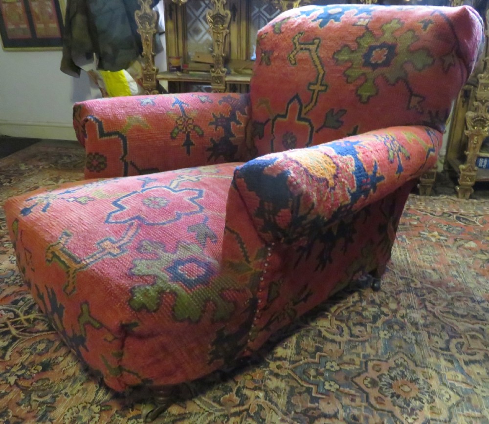 A low-slung, deep-seated Howard and Sons club armchair upholstered in a terracotta red and multi-coloured Ushak carpet pattern with turned legs on brass castors. View V