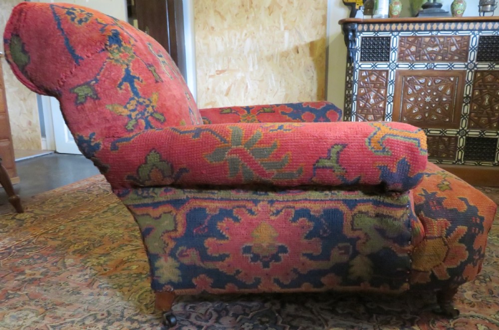 A low-slung, deep-seated Howard and Sons club armchair upholstered in a terracotta red and multi-coloured Ushak carpet pattern with turned legs on brass castors. View VII