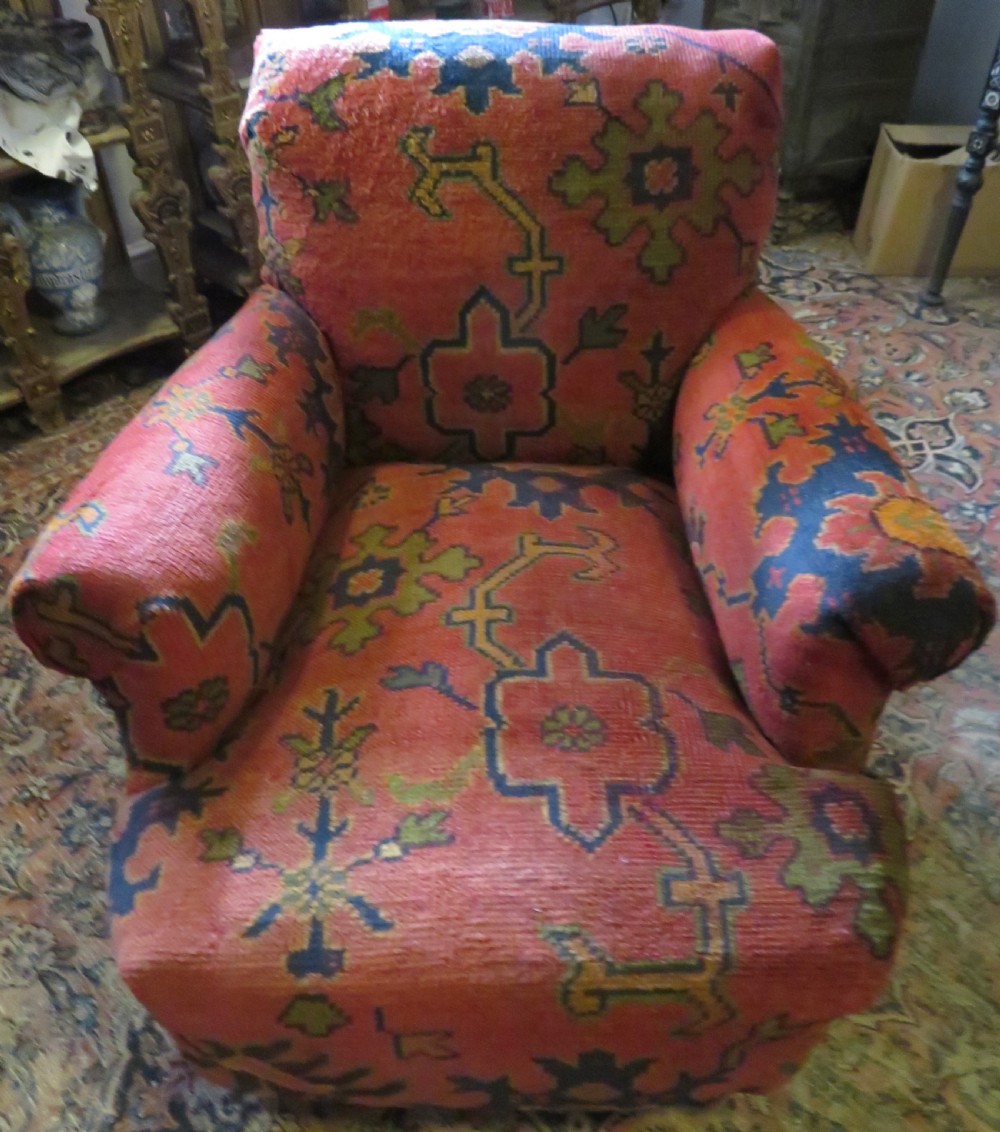 A low-slung, deep-seated Howard and Sons club armchair upholstered in a terracotta red and multi-coloured Ushak carpet pattern with turned legs on brass castors. View IX