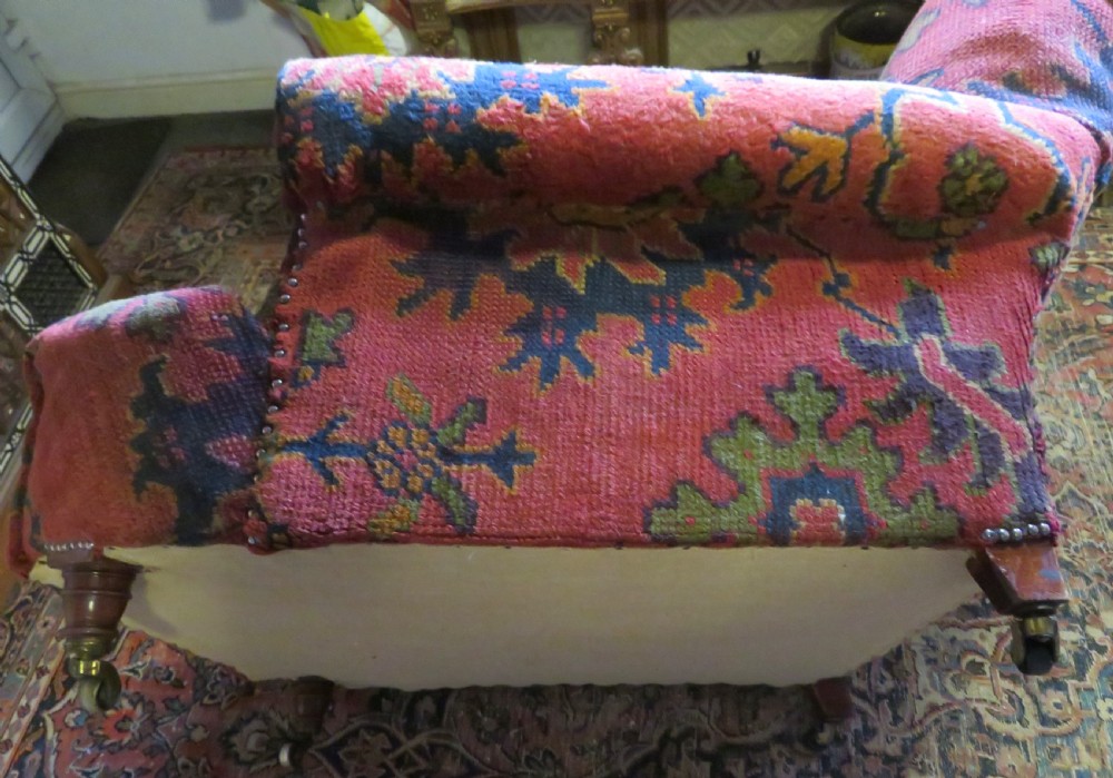 A low-slung, deep-seated Howard and Sons club armchair upholstered in a terracotta red and multi-coloured Ushak carpet pattern with turned legs on brass castors. View X