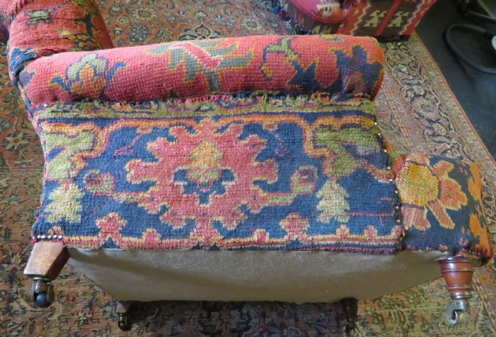 A low-slung, deep-seated Howard and Sons club armchair upholstered in a terracotta red and multi-coloured Ushak carpet pattern with turned legs on brass castors. View XI