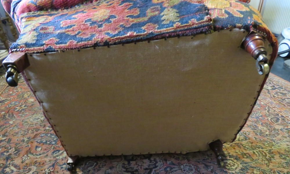 A low-slung, deep-seated Howard and Sons club armchair upholstered in a terracotta red and multi-coloured Ushak carpet pattern with turned legs on brass castors. View XV