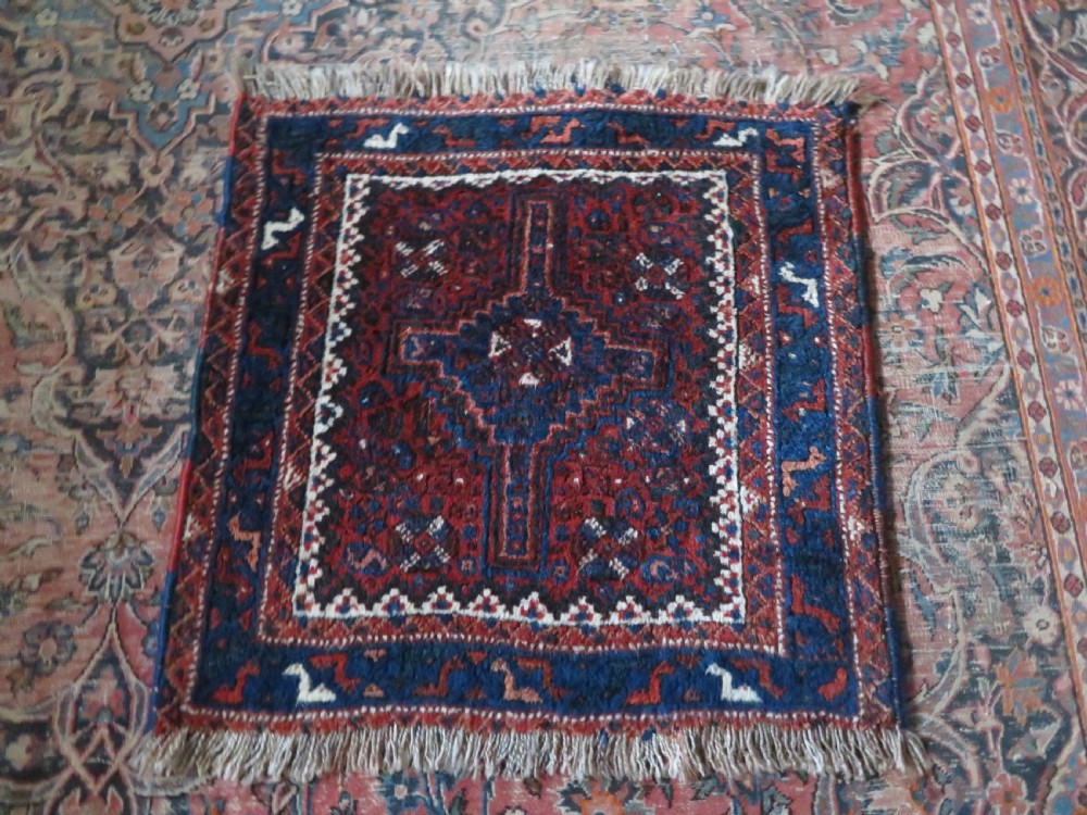An antique square hand-knotted tribal wool rug panel featuring a central stepped geometric medallion in red, indigo blue, and ivory with fringed edges. View X