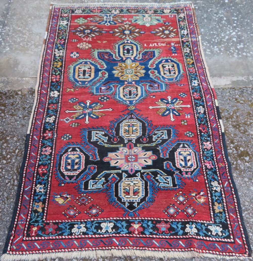 An antique Caucasian Karabagh handwoven wool rug featuring blue and black central geometric medallions on a red field with floral borders.