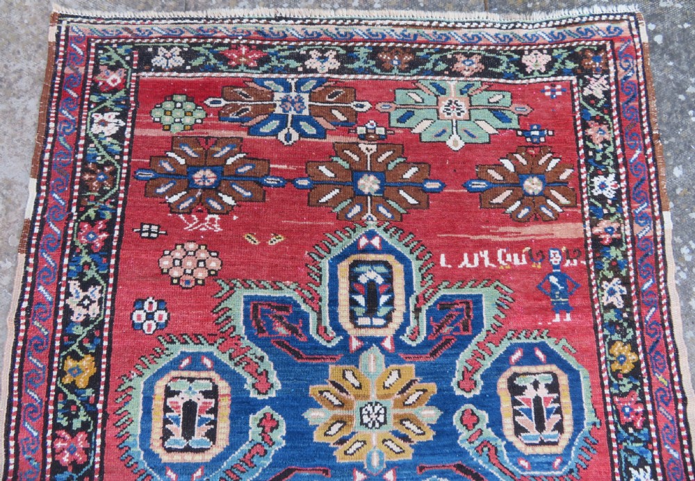An antique Caucasian Karabagh handwoven wool rug featuring blue and black central geometric medallions on a red field with floral borders. View II