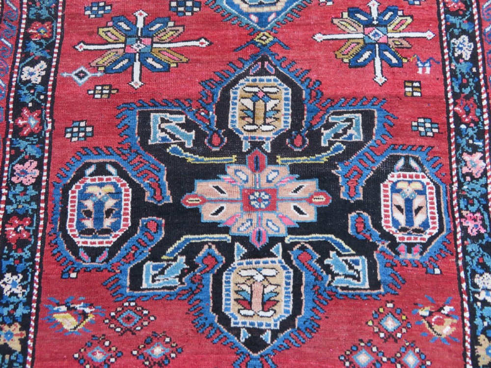 An antique Caucasian Karabagh handwoven wool rug featuring blue and black central geometric medallions on a red field with floral borders. View IV