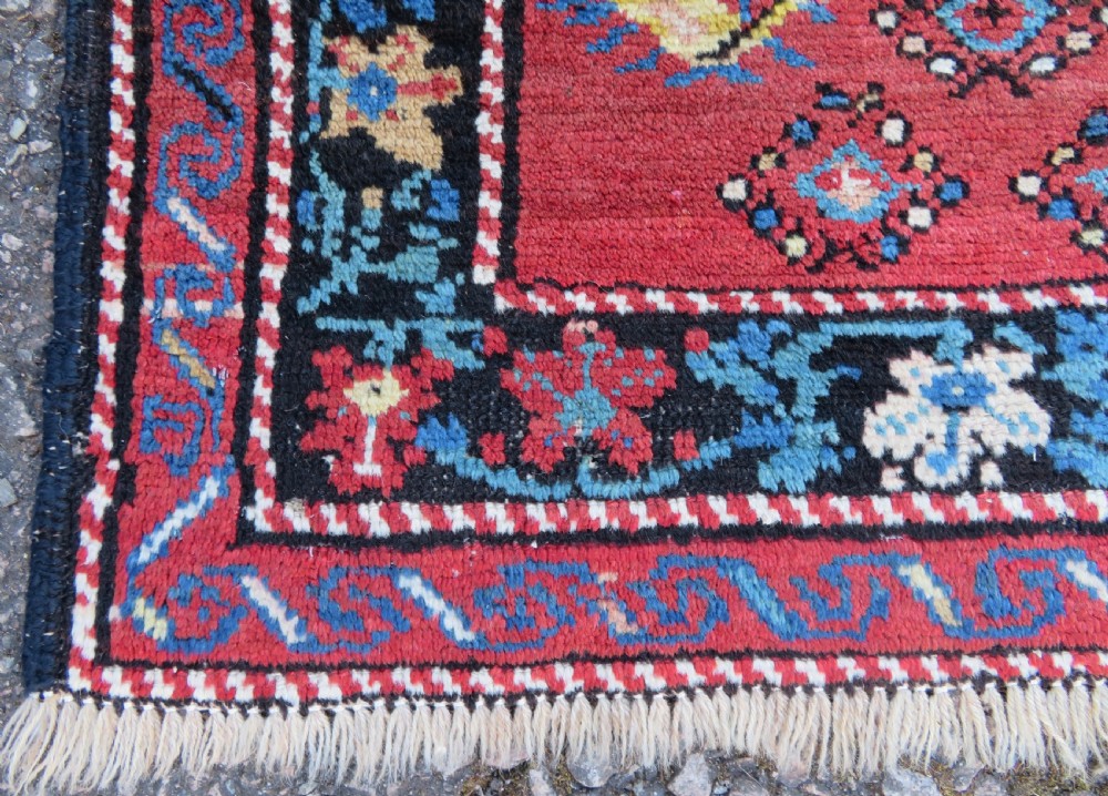 An antique Caucasian Karabagh handwoven wool rug featuring blue and black central geometric medallions on a red field with floral borders. View V