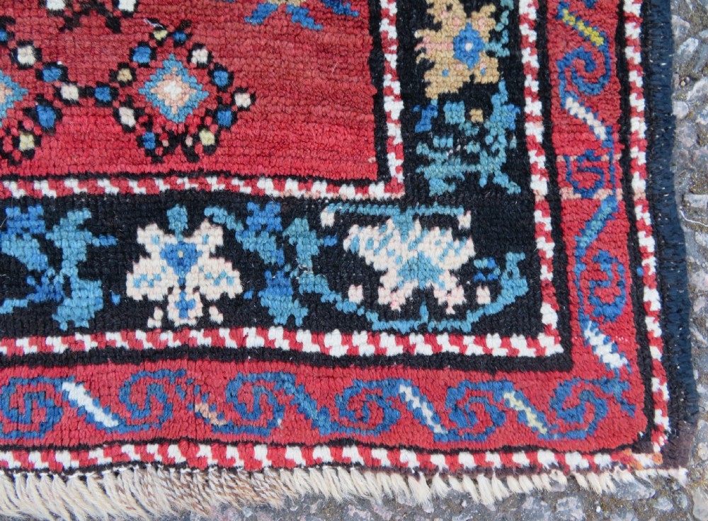 An antique Caucasian Karabagh handwoven wool rug featuring blue and black central geometric medallions on a red field with floral borders. View VI