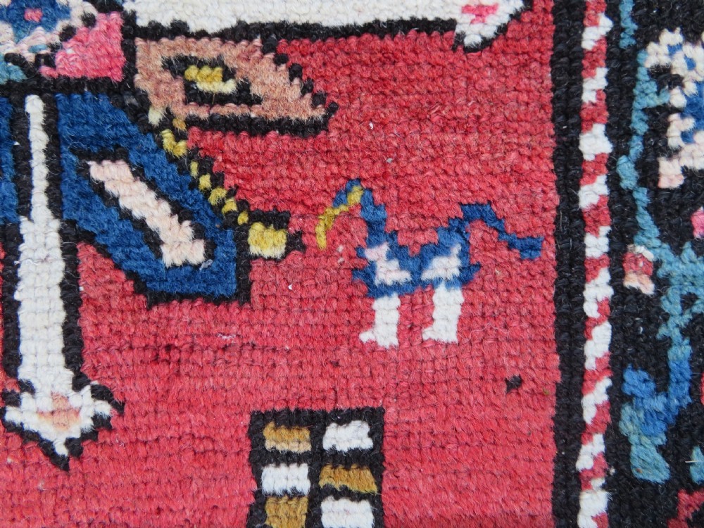 An antique Caucasian Karabagh handwoven wool rug featuring blue and black central geometric medallions on a red field with floral borders. View X