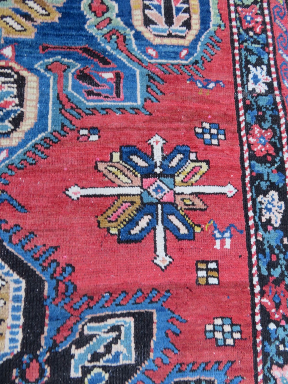 An antique Caucasian Karabagh handwoven wool rug featuring blue and black central geometric medallions on a red field with floral borders. View XIV