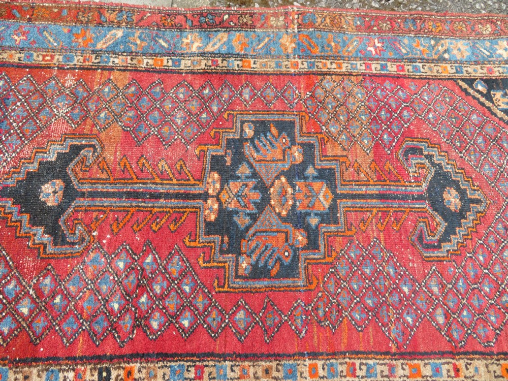 An antique hand-knotted Persian Sarab rug featuring a central dark blue geometric medallion on a red patterned field with light blue borders. View IV