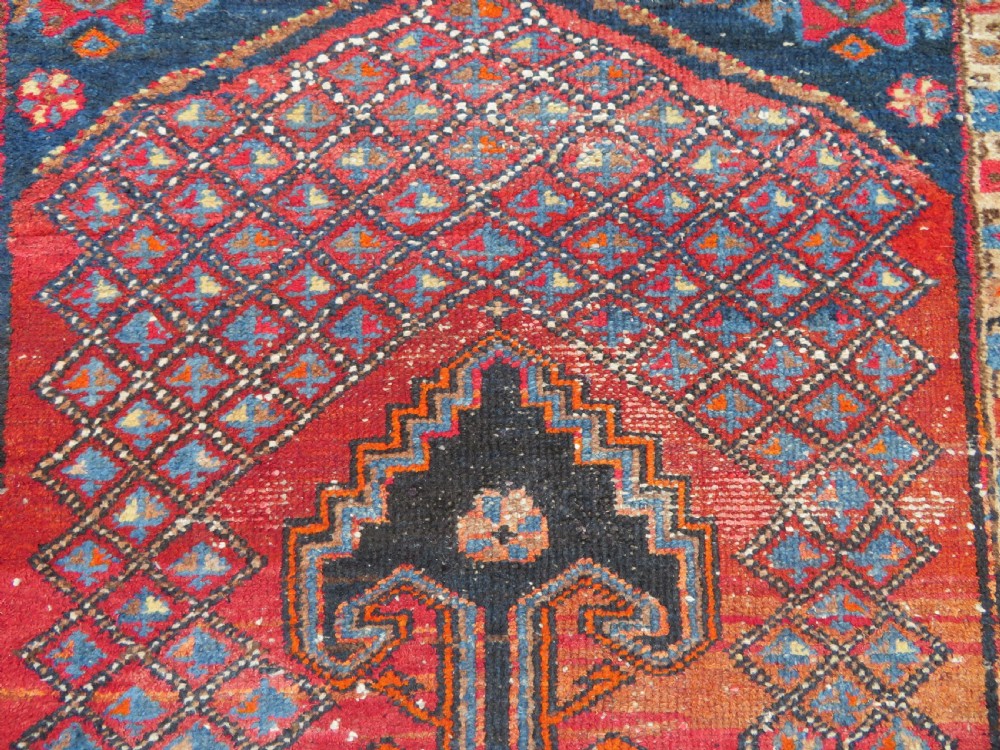 An antique hand-knotted Persian Sarab rug featuring a central dark blue geometric medallion on a red patterned field with light blue borders. View V