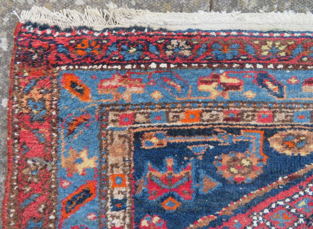 An antique hand-knotted Persian Sarab rug featuring a central dark blue geometric medallion on a red patterned field with light blue borders. View VI