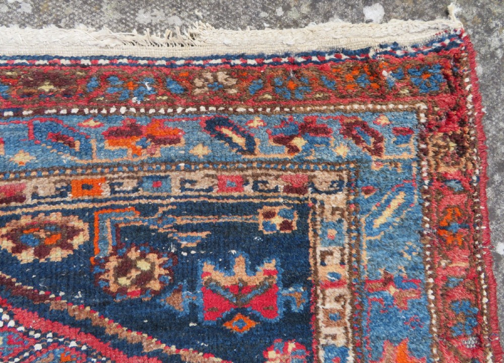 An antique hand-knotted Persian Sarab rug featuring a central dark blue geometric medallion on a red patterned field with light blue borders. View VII