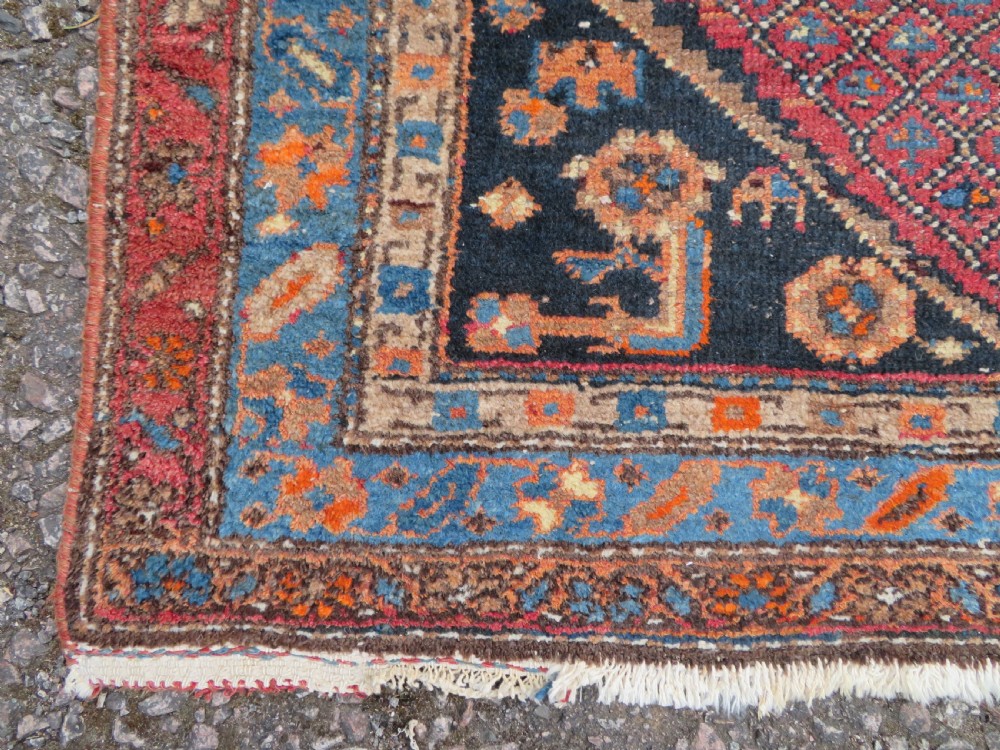 An antique hand-knotted Persian Sarab rug featuring a central dark blue geometric medallion on a red patterned field with light blue borders. View X
