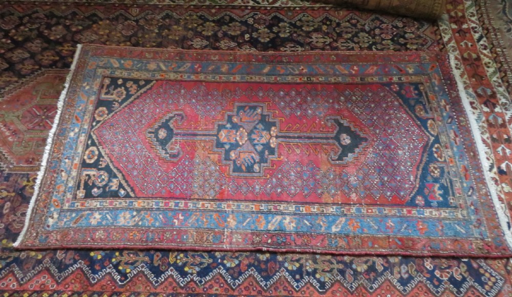 An antique hand-knotted Persian Sarab rug featuring a central dark blue geometric medallion on a red patterned field with light blue borders. View XVII