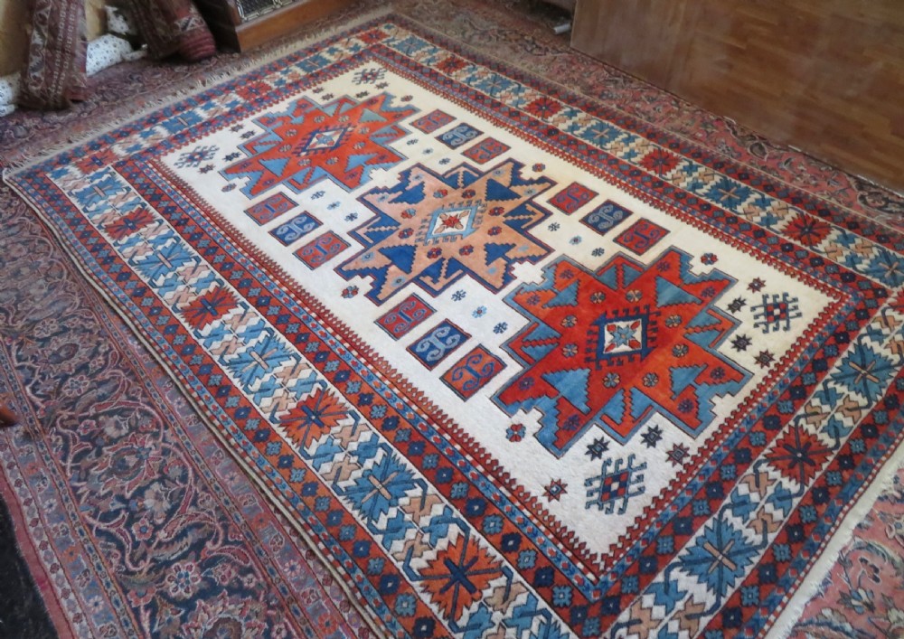 A hand-knotted wool carpet featuring three large geometric medallions in red, blue, and terracotta on a cream field, framed by multi-layered geometric borders. View III