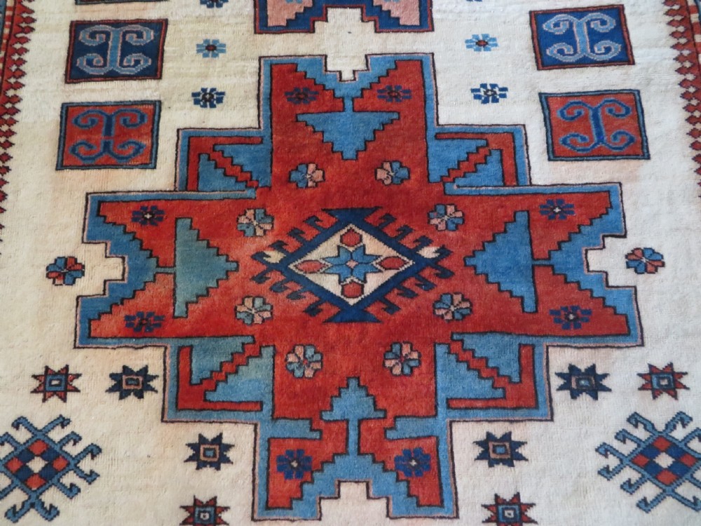 A hand-knotted wool carpet featuring three large geometric medallions in red, blue, and terracotta on a cream field, framed by multi-layered geometric borders. View IV