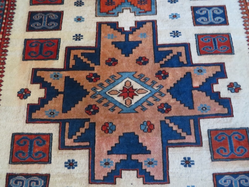 A hand-knotted wool carpet featuring three large geometric medallions in red, blue, and terracotta on a cream field, framed by multi-layered geometric borders. View V