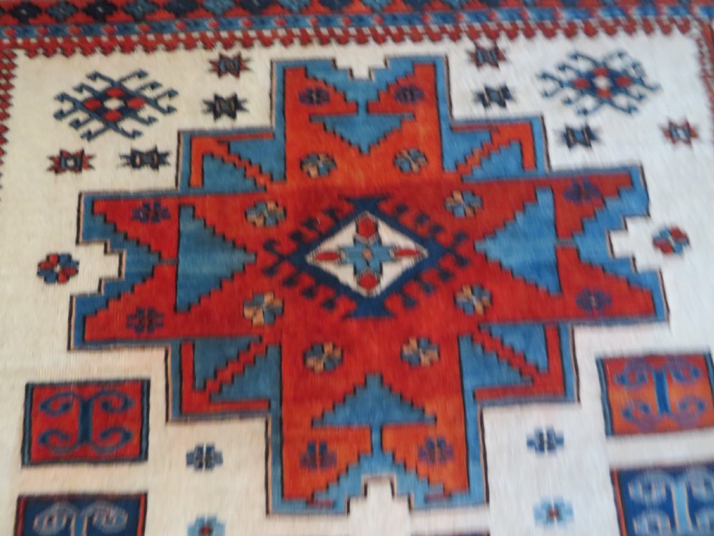 A hand-knotted wool carpet featuring three large geometric medallions in red, blue, and terracotta on a cream field, framed by multi-layered geometric borders. View VI