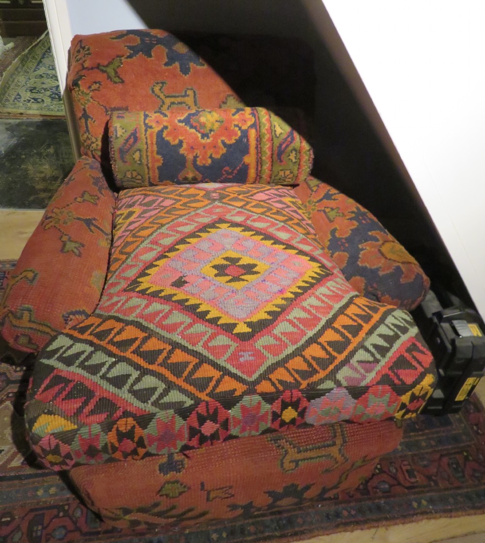A low-slung, deep-seated Howard and Sons club armchair upholstered in a terracotta red and multi-coloured Ushak carpet pattern with turned legs on brass castors. View XXIII