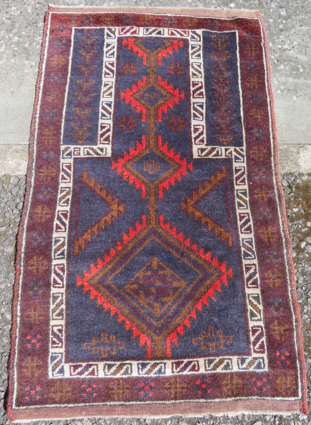 A hand-knotted antique Persian Baluch prayer rug with geometric diamond motifs in rust red and brown against a dark blue central field.