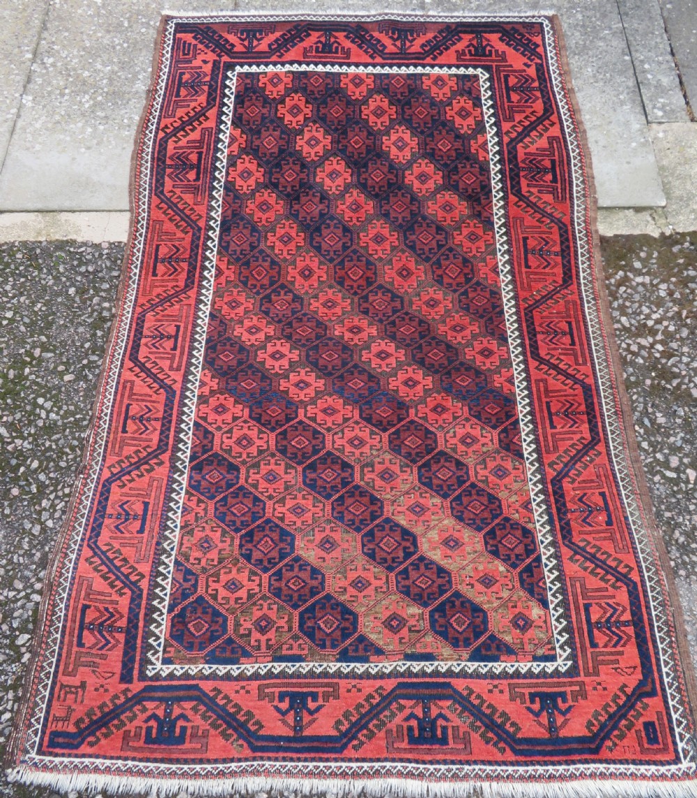 An antique hand-knotted Baluch wool rug featuring repeating geometric diamond motifs in deep red, navy blue, and brown tones with fringed ends.
