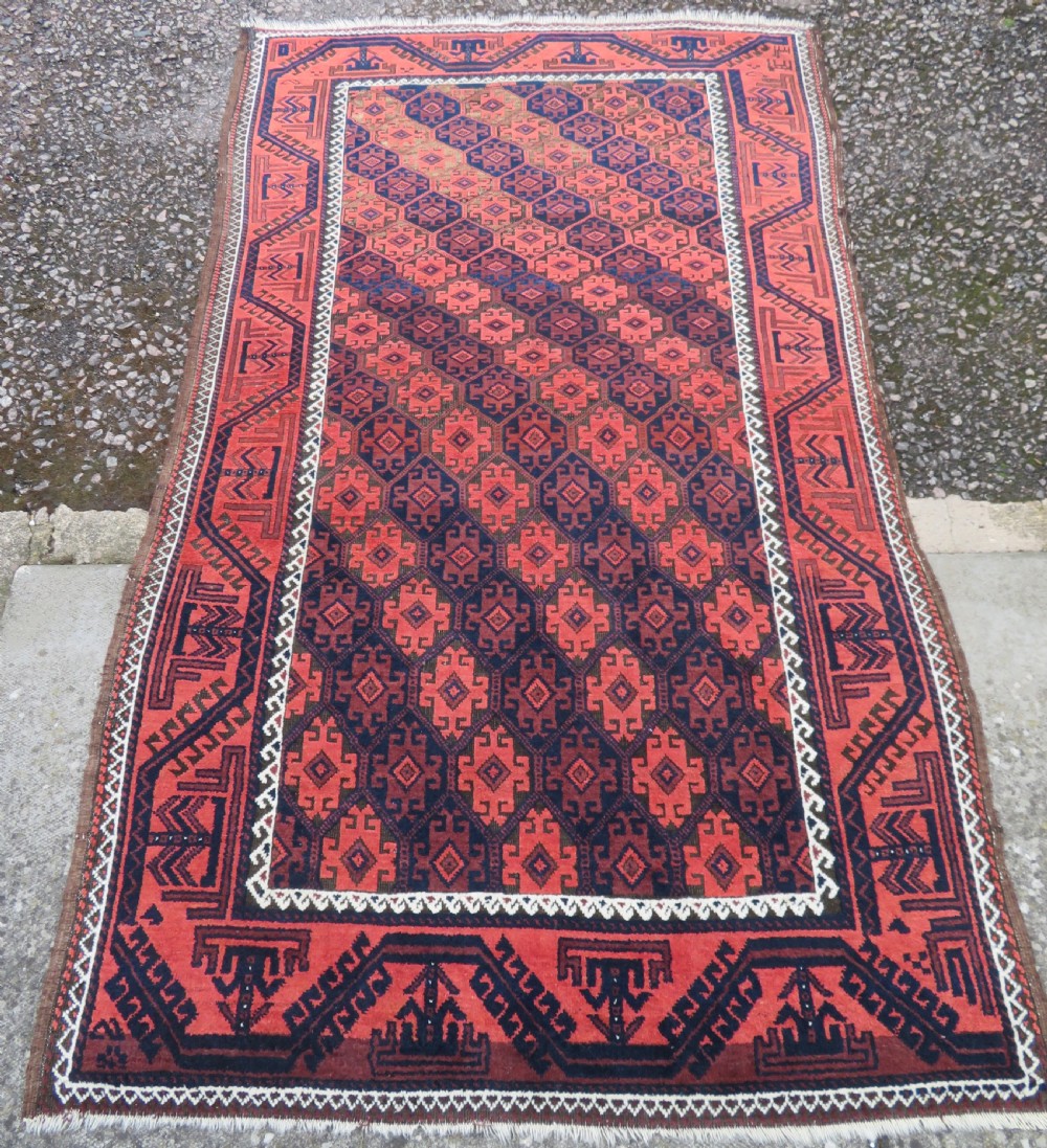 An antique hand-knotted Baluch wool rug featuring repeating geometric diamond motifs in deep red, navy blue, and brown tones with fringed ends. View II