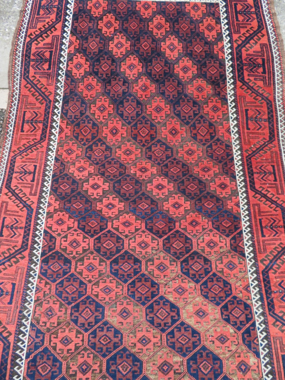 An antique hand-knotted Baluch wool rug featuring repeating geometric diamond motifs in deep red, navy blue, and brown tones with fringed ends. View III