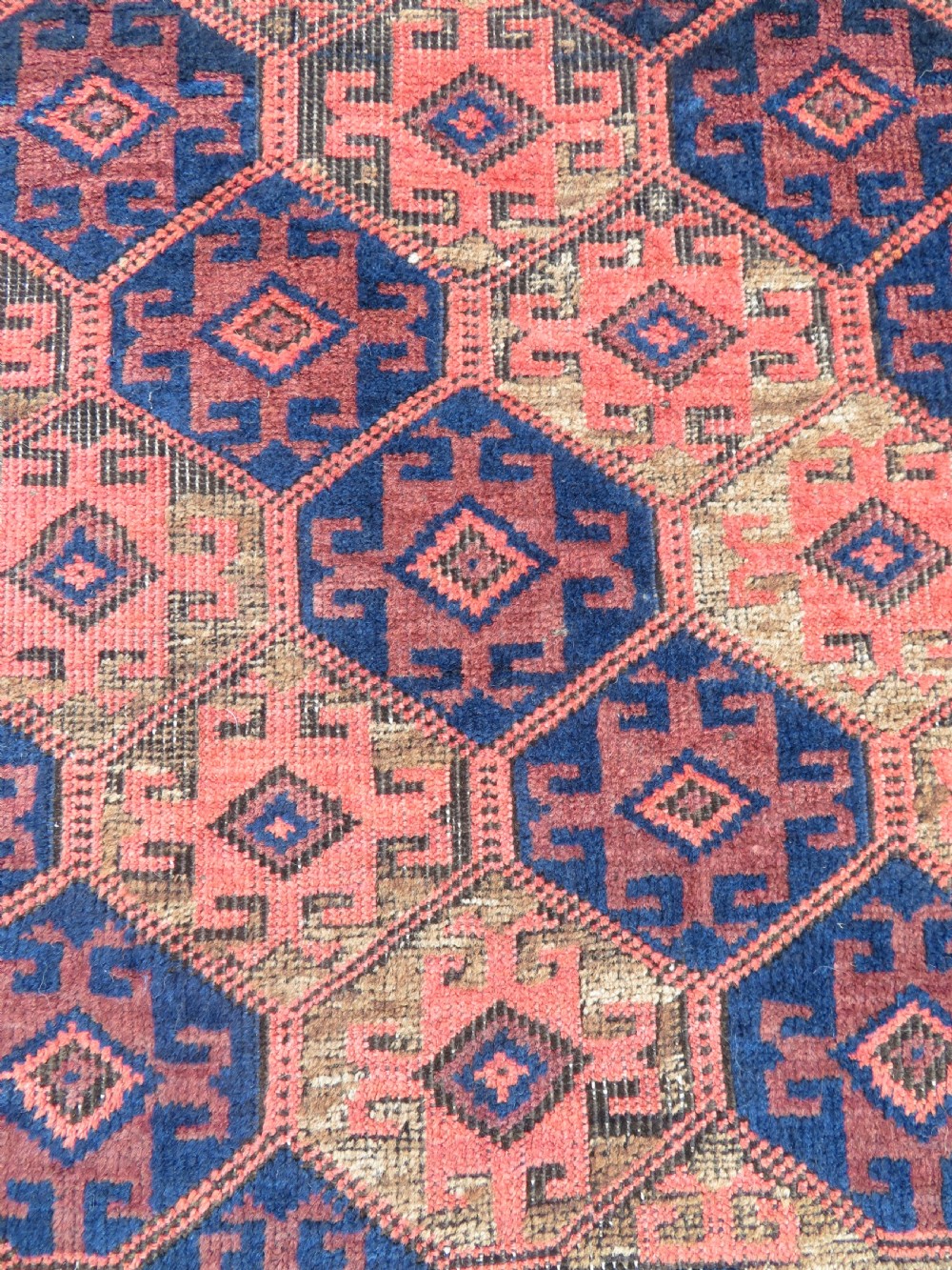 An antique hand-knotted Baluch wool rug featuring repeating geometric diamond motifs in deep red, navy blue, and brown tones with fringed ends. View IV