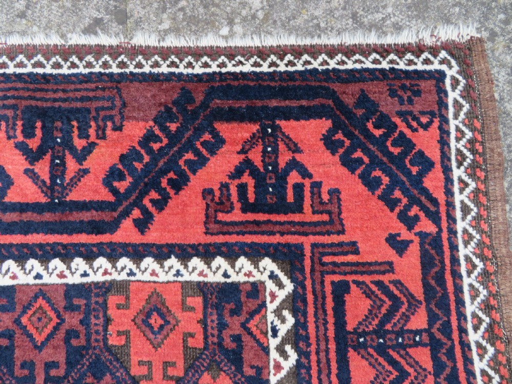 An antique hand-knotted Baluch wool rug featuring repeating geometric diamond motifs in deep red, navy blue, and brown tones with fringed ends. View V