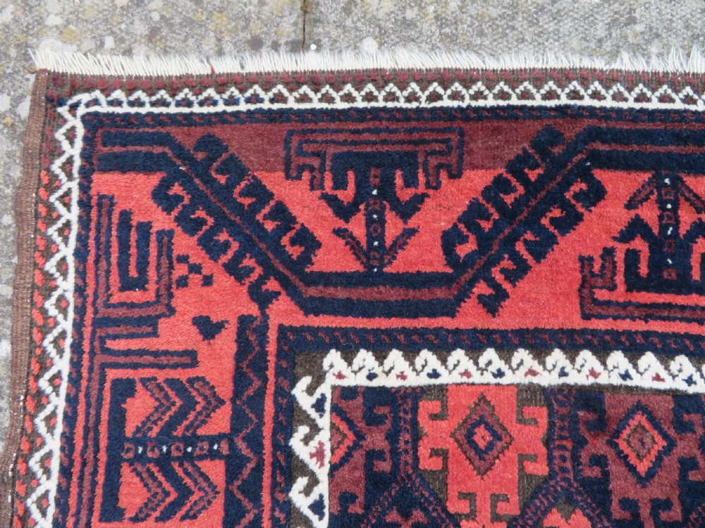 An antique hand-knotted Baluch wool rug featuring repeating geometric diamond motifs in deep red, navy blue, and brown tones with fringed ends. View VI
