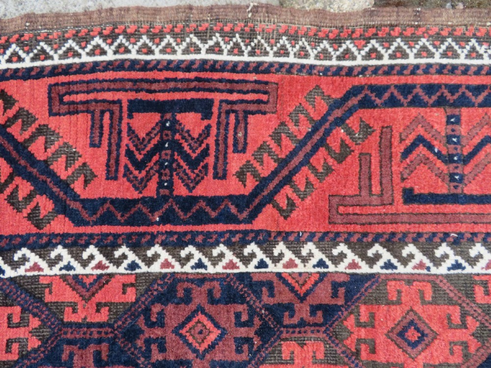 An antique hand-knotted Baluch wool rug featuring repeating geometric diamond motifs in deep red, navy blue, and brown tones with fringed ends. View X