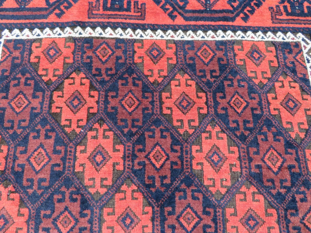 An antique hand-knotted Baluch wool rug featuring repeating geometric diamond motifs in deep red, navy blue, and brown tones with fringed ends. View XI