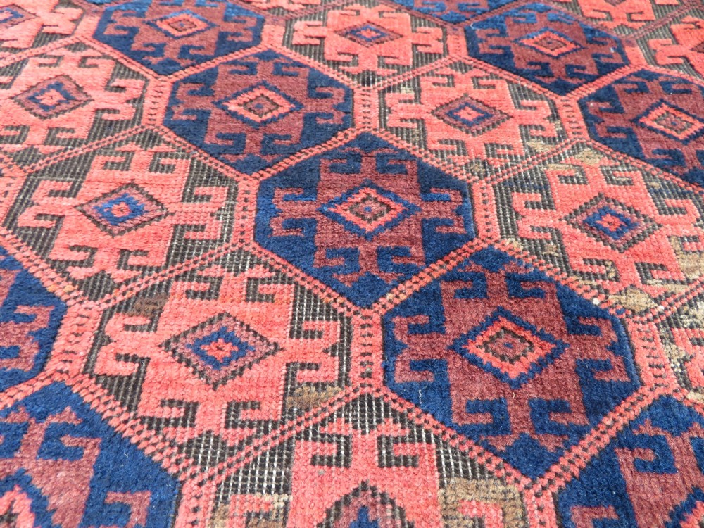 An antique hand-knotted Baluch wool rug featuring repeating geometric diamond motifs in deep red, navy blue, and brown tones with fringed ends. View XII
