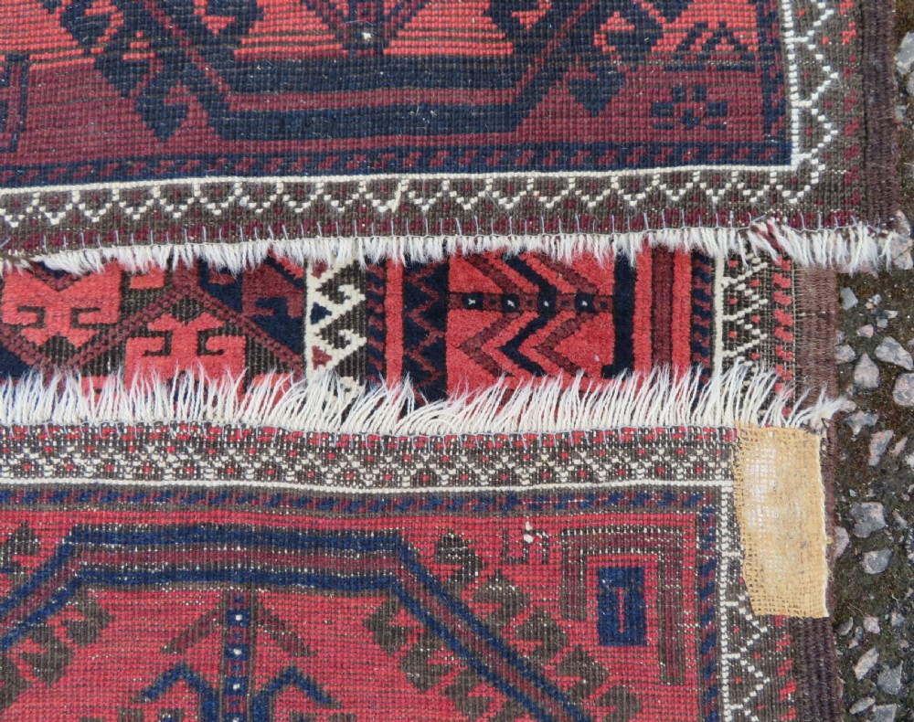 An antique hand-knotted Baluch wool rug featuring repeating geometric diamond motifs in deep red, navy blue, and brown tones with fringed ends. View XIV